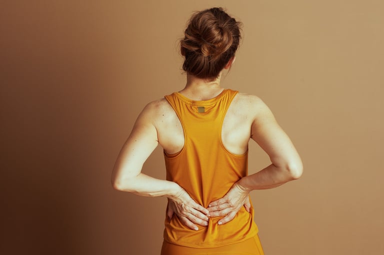 Mujer con top atlético amarillo-naranja presionando su espalda baja con las manos, expresando dolor y tensión física